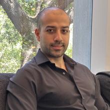 Khashayar Moshksayan Khashayar Moshksayan sits on a black couch, wearing a brown button-up shirt. Behind him is a white wall with a window. Through the window are bright green leaves and the thick trunk and branches of a tree.