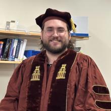 Keegan Lorenz-Ochoa Keegan Lorenz-Ochoa wears the doctoral regalia for the University of Texas at Austin. The regalia is dark red, with a brown velvet sash around the neck which has the tower embroidered along both sides, in gold. He also wears a dark red velvet cap with a gold tassel, and glasses. Lorenz-Ochoa stands in front of a bookshelf of chemistry books, in a white room.