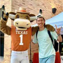 Yanbing Zhou Yanbing Zhou wears a teal t-shirt, black backpack, light blue shorts, and a beige baseball cap turned backwards. Next to him is Hook'em, a mascot of a brown bull with white and black horns, an orange Texas jersey, white shorts, and a beige cowboy hat. Both Hook'em and Yanbing smile and make the "Hook 'em" hand sign in front of a beige brick building. Behind them is a light blue tent over a table draped in red cloth.
