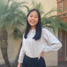 Lisa Wang Lisa Wang wears a grey long-sleeved blouse and a black skirt. She smiles with one hand on her hip in front of a beige wall with two small palm trees behind her.